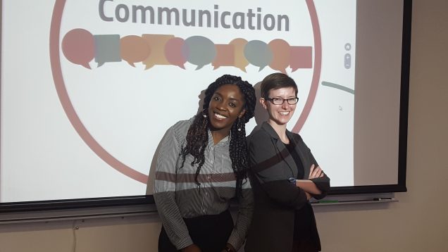 Two people stand in front of a slideshow that reads "Communication"