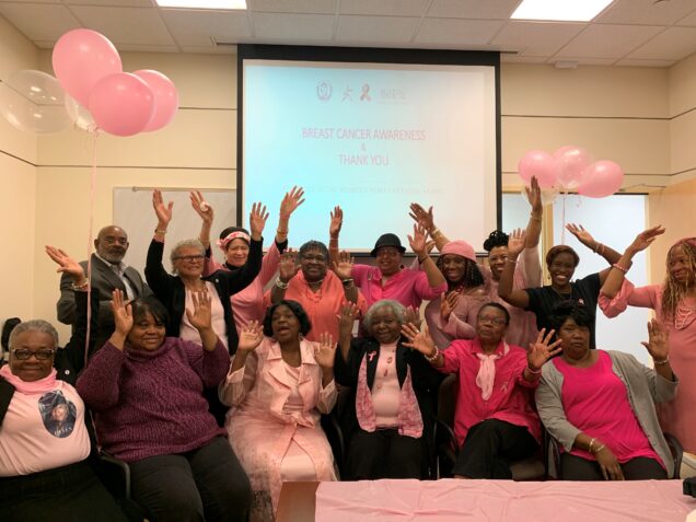 A large group of Black women stand with their arms up in celebration in front of a screen that says "Breast Cancer Awareness and Thank You"