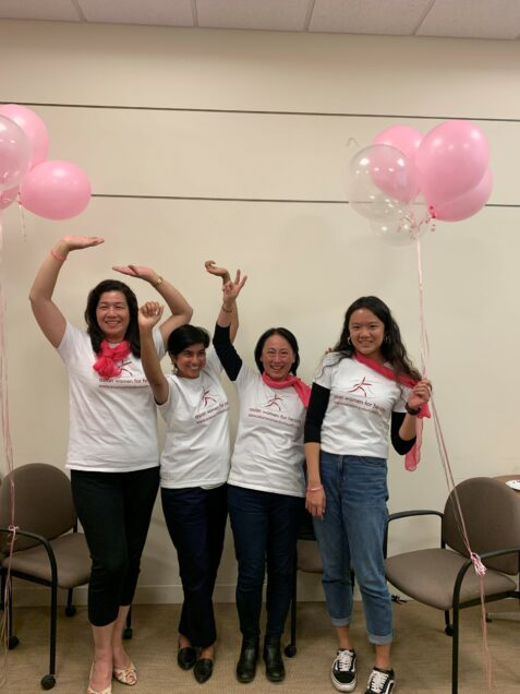 Four Asian women stand with their arms up in celebration with pink balloons.
