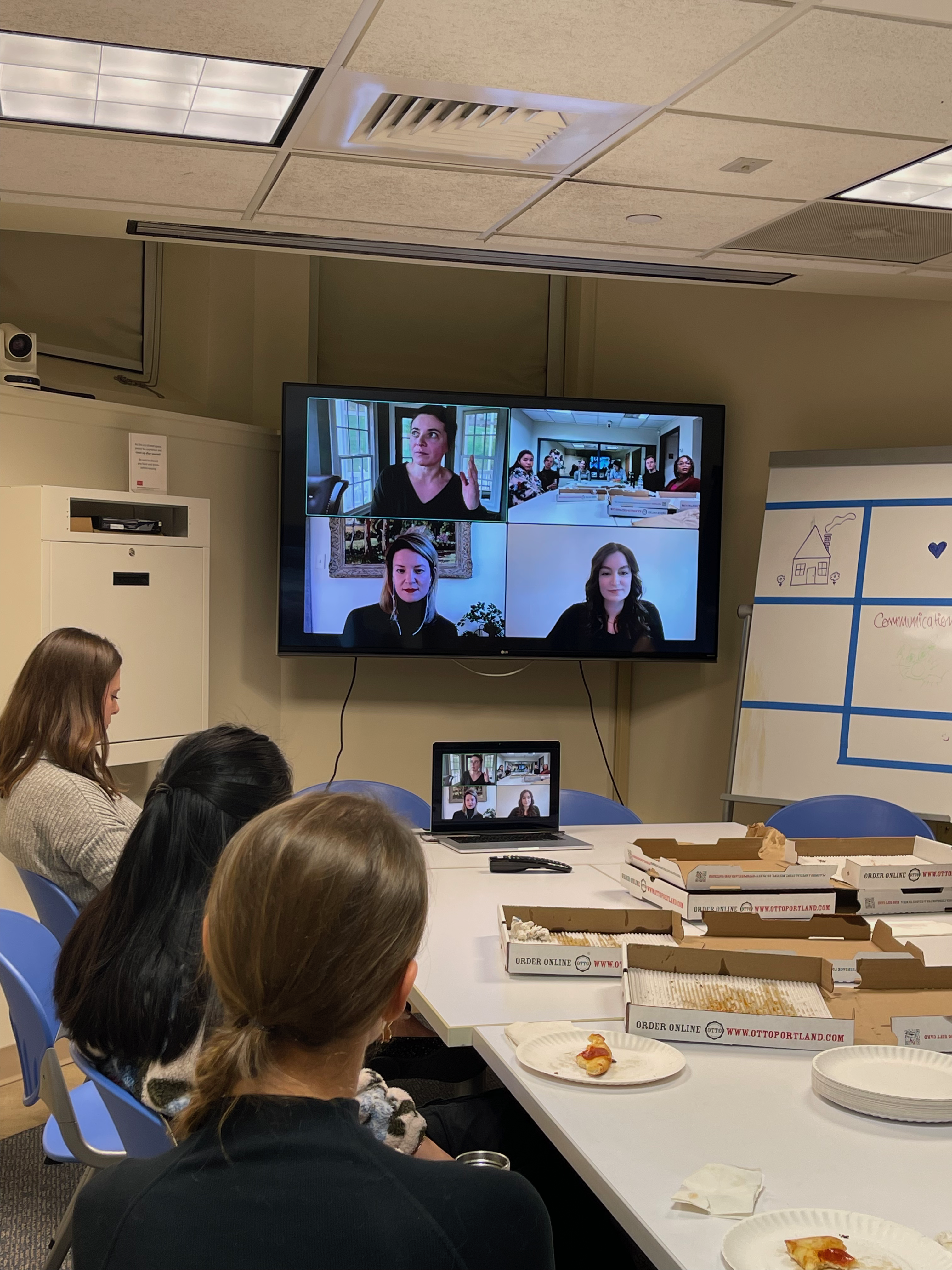 An image of students facing a TV screen, half eaten pizza slices on paper plates on the table in front of them, with a Zoom video on display.