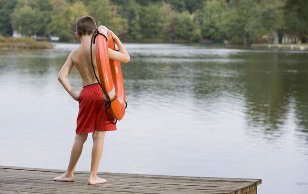 Boy with life preserver standing on pier by lake.