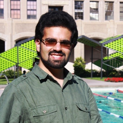 A man with black hair, a beard, sunglasses, and a green button up shirt smiles in front of an outdoor swimming pool