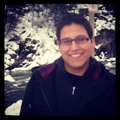 A man with black hair, glasses, and a black coat is smiling in front of a snowy mountain