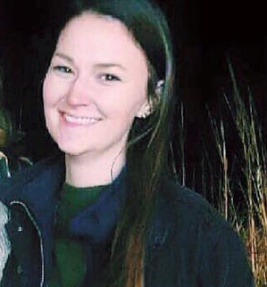 A woman with brown hair, a green shirt, and a black coat smiles outdoors