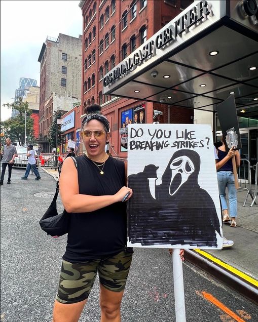 A woman poses with a sign on a NYC street. Sign image: the "Scream" figure, text "Do you like breaking strikes?"