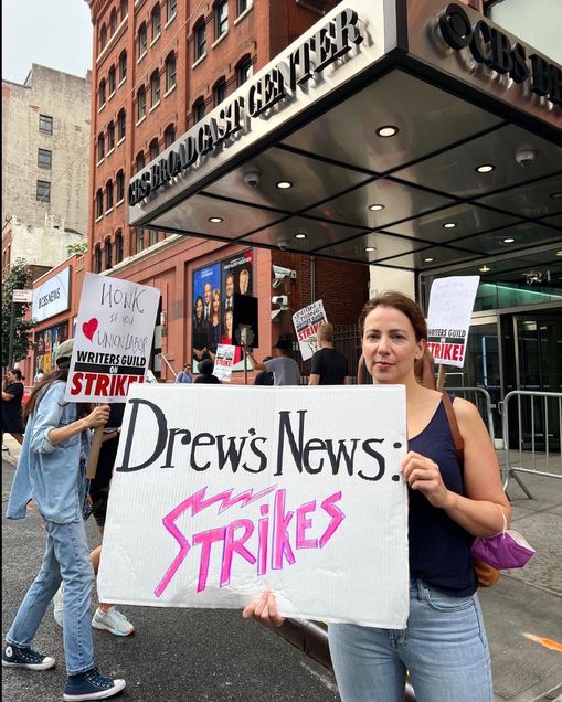 A woman poses with a sign on an NYC street. Sign text: "Drew's News: Strikes."