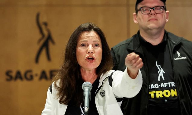 A woman (Fran Drescher) stands behind a microphone and points. A man stands behind her at right. Behind them is a logo on the wall that says SAG-AFTRA.