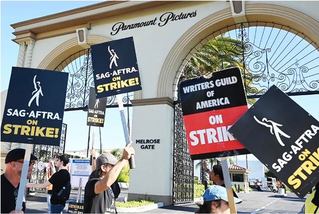 Four people march in a line in front of an arched entrance to the studio Paramount Pictures. Signs say "SAG-Aftra on Strike."