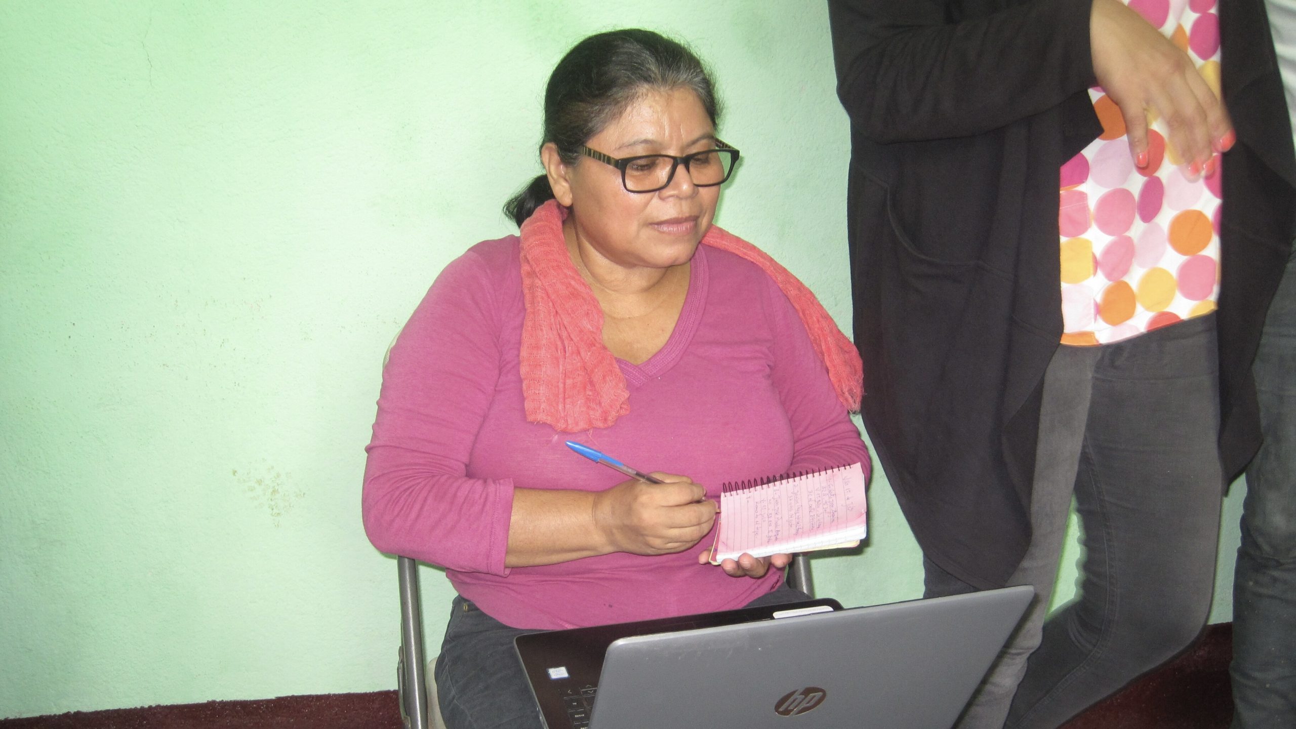 picture of Damaris López holding a computer in her lap, against a green plan wall.