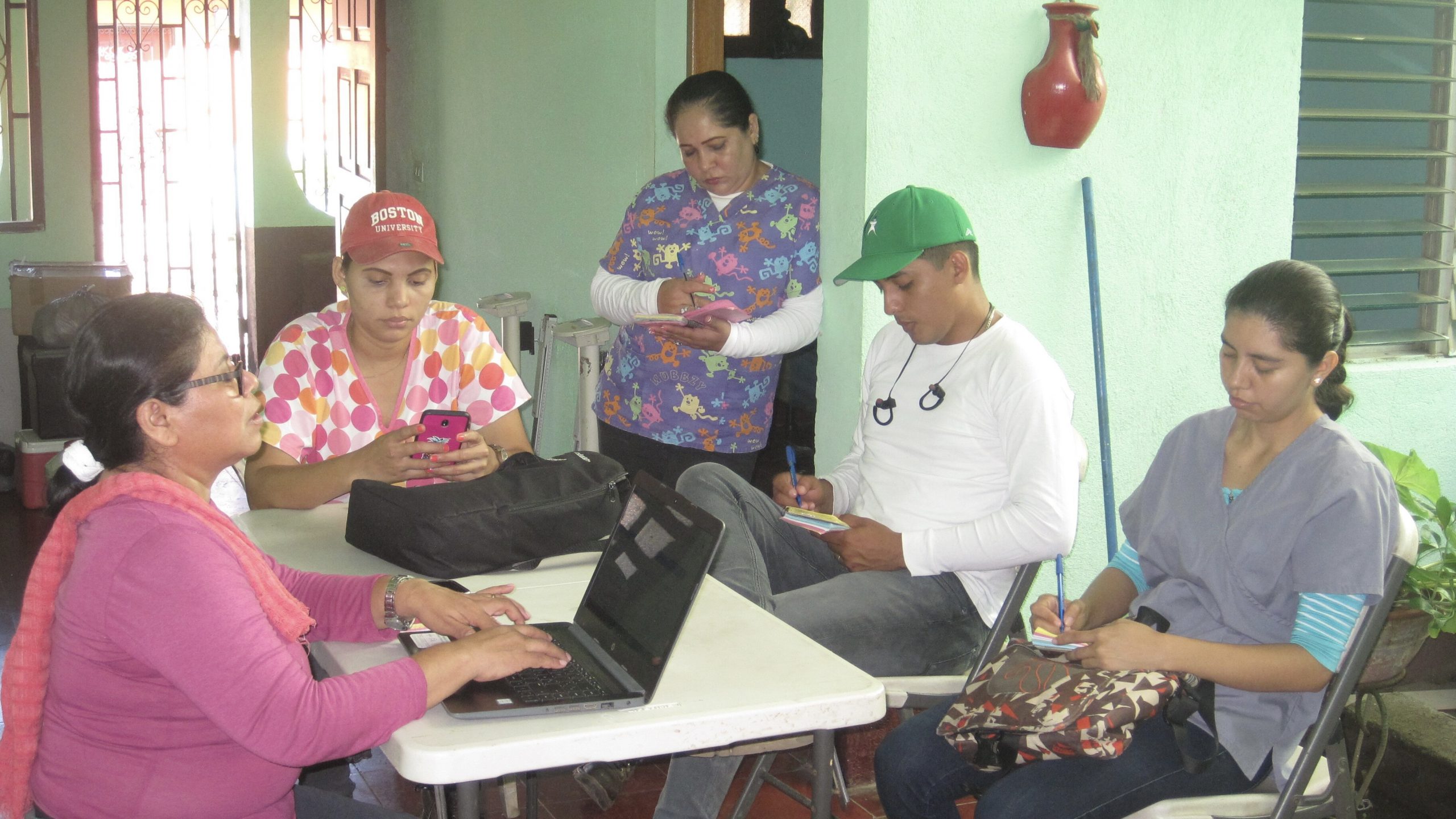 a picture of Nicaraguan Team. woman on the left is typing on a computer, the woman next to her is looking at her phone, the next woman is standing while taking notes, the men next to her is sitting down while taking notes, the woman next to him is sitting down while taking notes. they are all around a table.
