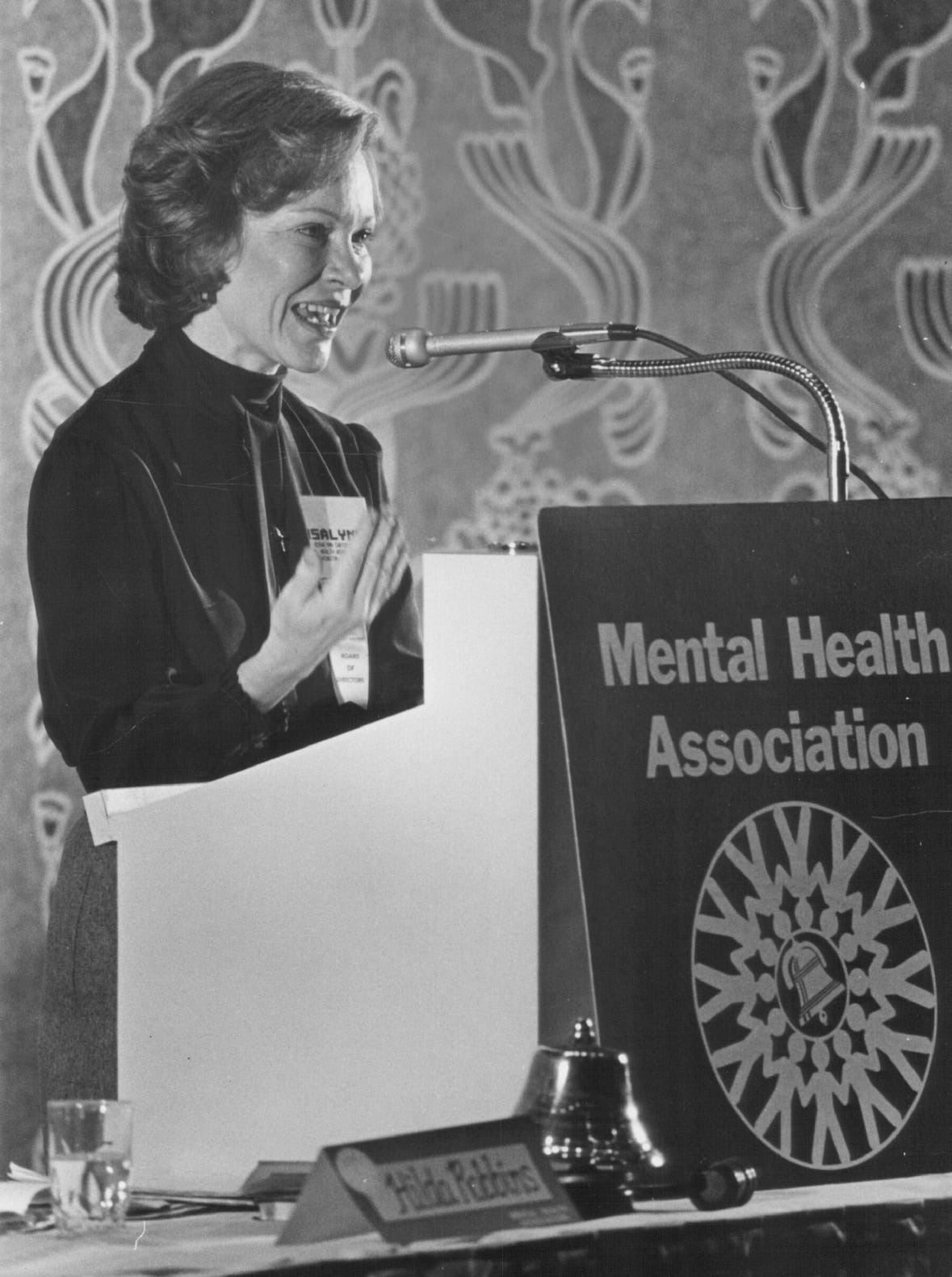 A black and white photo of Mrs. Rosalynn Carter speaking at a podium in front of a poster titled 'Mental Health Association'.