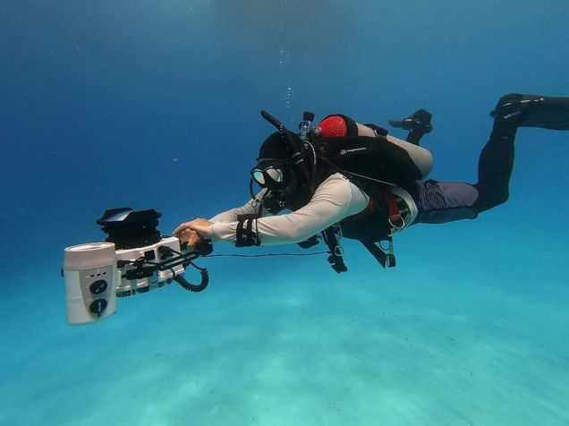 Underwater photo of Fabio Buitrago, dressed in full scuba diving gear, swimming in light blue water while holding a large underwater camera appratus.