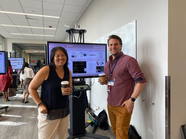 Dr Ko and Dr Dries stand in front of a research poster presented on a tv screen 