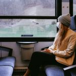 Person wearing a mask and a beanie sitting on a train, looking out the window at a rural landscape.