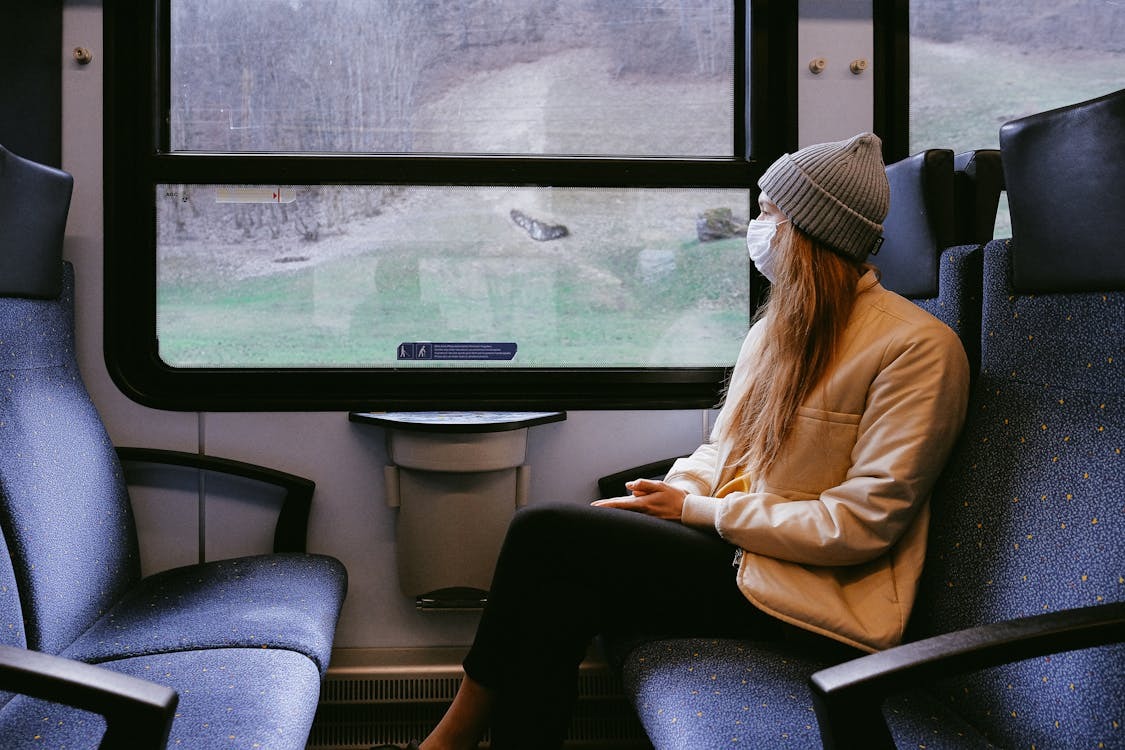 Person wearing a mask and a beanie sitting on a train, looking out the window at a rural landscape.