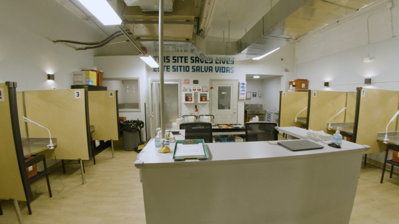 Interior of an overdose prevention center with individual numbered booths, harm reduction supplies, and a sign reading 'This Site Saves Lives' in English and Spanish.