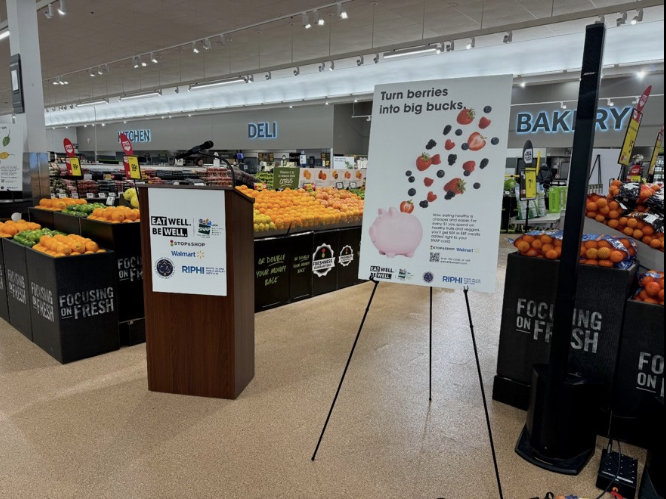 A podium in a grocery store promotes a health campaign with a sign reading "Turn berries into big bucks," showing berries falling into a piggy bank. Fresh produce displays are visible in the background.