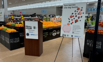 A podium in a grocery store promotes a health campaign with a sign reading "Turn berries into big bucks," showing berries falling into a piggy bank. Fresh produce displays are visible in the background.