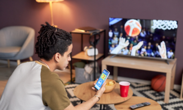 A person sitting in a living room holds a smartphone with a sports betting app open while watching a basketball game on TV