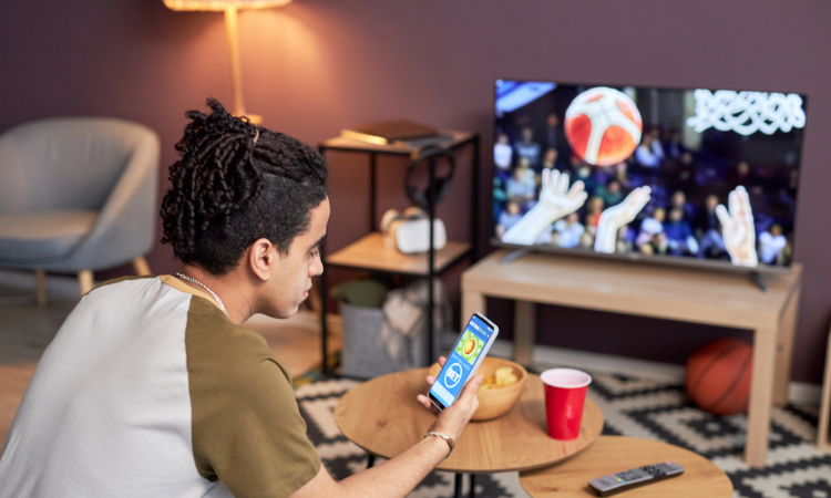 A person sitting in a living room holds a smartphone with a sports betting app open while watching a basketball game on TV