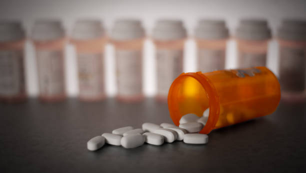 Orange prescription bottle tipped over with white pills spilling out, with several blurred prescription bottles in the background.