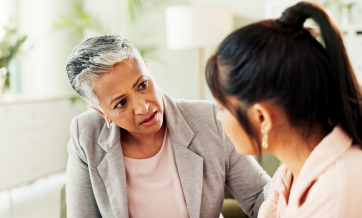 An older woman speaks attentively with a younger woman in a quiet, well-lit office or counseling setting.