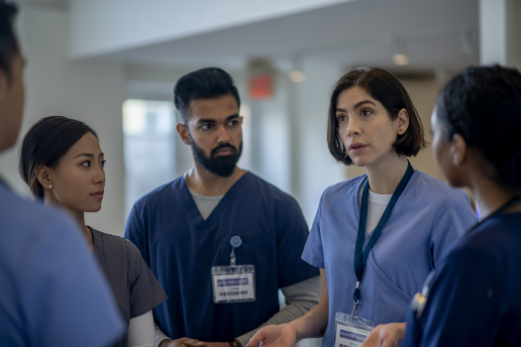 A group of four healthcare workers in scrubs and ID badges having a serious discussion in a hospital hallway.