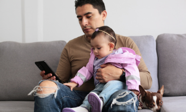 A father sits on a gray couch holding a baby in a pink jacket on his lap while looking at his phone, with a small brown and white dog resting beside him.