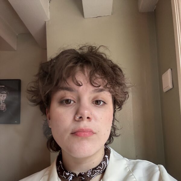 Photo of a person looking at the camera with short brown hair, brown eyes, and light peach skin. They have 2 nose rings, are wearing a brown bandana, and a button down cream-color shirt. The background is taupe.