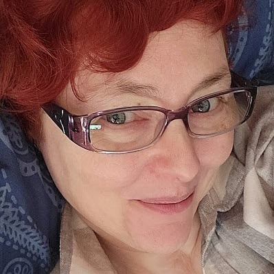 Photo of a smiling person with bright red hair, translucent purple glasses, blue eyes, and light peach color skin. They are wearing a grey sweatshirt and their head is resting on a blue and light blue patterned fabric.