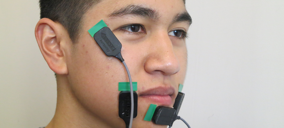  In front of a gray wall, a man with dark hair and dark eyes has four small black and green sensors placed on his face in various places – on both sides of his mouth, on his chin, and beside his eye