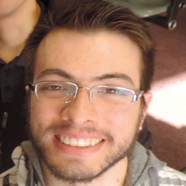 A man with brown hair, a brown beard, glasses, and a gray shirt is smiling in front of a brown background