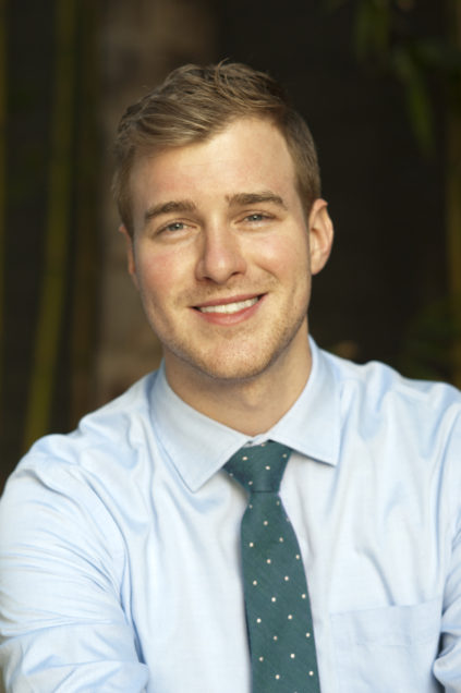 A man with brown hair, a light blue shirt, and a green spotted tie is smiling in front of a brown background